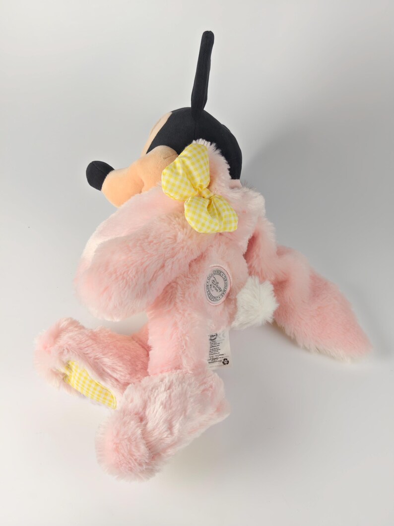 May include: A plush toy featuring a pink bunny costume with a yellow gingham bow and ears. The toy has a black and tan head, resembling a cartoon character. The bunny's feet and ears have yellow gingham accents. A white tag is attached.