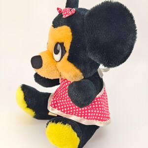 May include: A vintage plush toy of a cartoon character, featuring black fur, yellow face and feet, and a red polka dot dress. The character has large black ears, a small red bow, and white eyes. The toy is sitting.