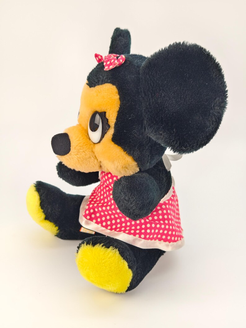 May include: A vintage plush toy of a cartoon character, featuring black fur, yellow face and feet, and a red polka dot dress. The character has large black ears, a small red bow, and white eyes. The toy is sitting.