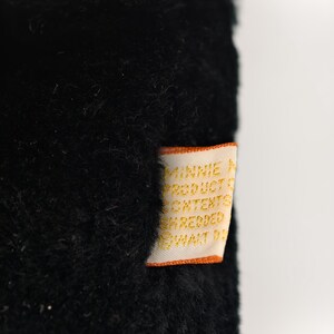 May include: Close-up of a black plush toy with a tag. The tag is orange and white with text that reads "MINNIE PRODUCT CONTENTS SHREDDED WALT D". The plush toy has a soft, fuzzy texture.