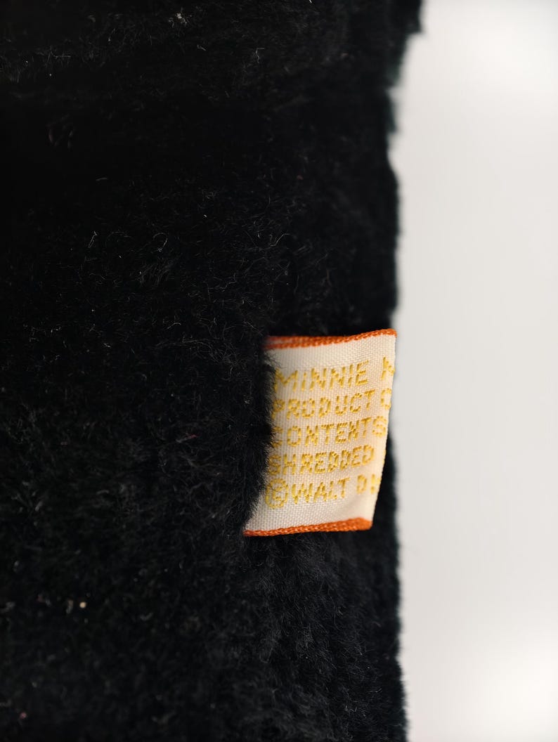 May include: Close-up of a black plush toy with a tag. The tag is orange and white with text that reads "MINNIE PRODUCT CONTENTS SHREDDED WALT D". The plush toy has a soft, fuzzy texture.