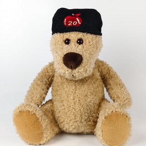 May include: A soft toy teddy bear with light brown fur, a dark brown nose, and dark eyes. The bear is wearing a black cap with a red apple and the number "20" embroidered on it. The bear is sitting on a white surface.