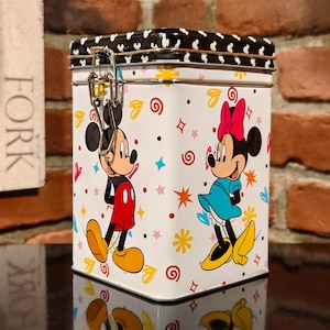 Op de afbeelding: Een witte rechthoekige blikken doos met een zwart stippen deksel en een metalen sluiting. De doos toont Mickey en Minnie Mouse stripfiguren, met kleurrijke bloemen- en sterontwerpen.