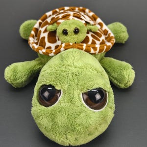 Puede incluir: Un juguete de peluche con forma de tortuga que presenta un cuerpo verde suave con grandes ojos de color marrón oscuro y una tortuga verde más pequeña unida a su espalda. El caparazón de la tortuga grande tiene un patrón marrón y blanco a juego.