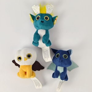 以下が含まれることがあります： 3つのぬいぐるみファンタジー生き物：黄色い髪と銀色のアクセントのターコイズのゴブリン、茶色の翼を持つ黄色と白のフクロウ、水色の翼を持つ青いドラゴン。それぞれ大きな丸い目とタグが付いています。