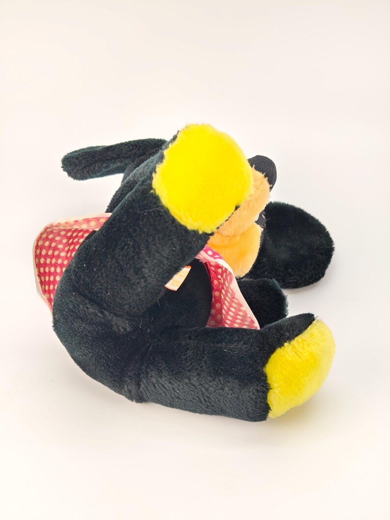May include: A plush toy dog with black fur, yellow paws, and a peach-colored face. The dog is wearing a red polka-dot dress. The toy is lying on its side against a white background.