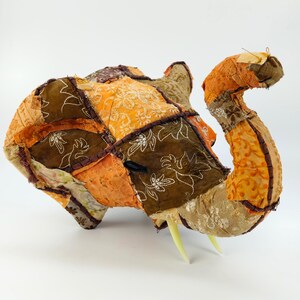 Könnte beinhalten: Handgefertigte Elefantenskulptur aus Patchwork-Stoff in Orange-, Braun- und Beigetönen. Der Elefant hat einen Rüssel, Stoßzähne und detaillierte Nähte. Das Patchwork-Design zeigt florale Muster und geometrische Formen.