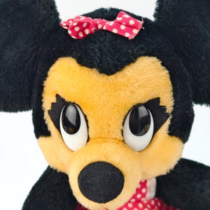 May include: A plush toy of a cartoon mouse with black fur, a yellow face, and large, expressive eyes. The mouse wears a red bow with white polka dots and a red dress with white polka dots. The toy has large black ears.