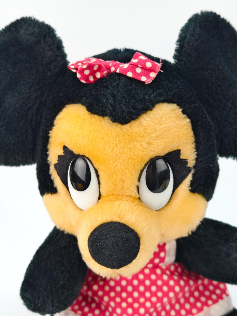 May include: A plush toy of a cartoon mouse with black fur, a yellow face, and large, expressive eyes. The mouse wears a red bow with white polka dots and a red dress with white polka dots. The toy has large black ears.