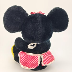 May include: A plush black mouse toy with large ears, a red polka dot bow, and a pink polka dot apron. The toy is sitting down, with its back facing the viewer. The apron has white trim.