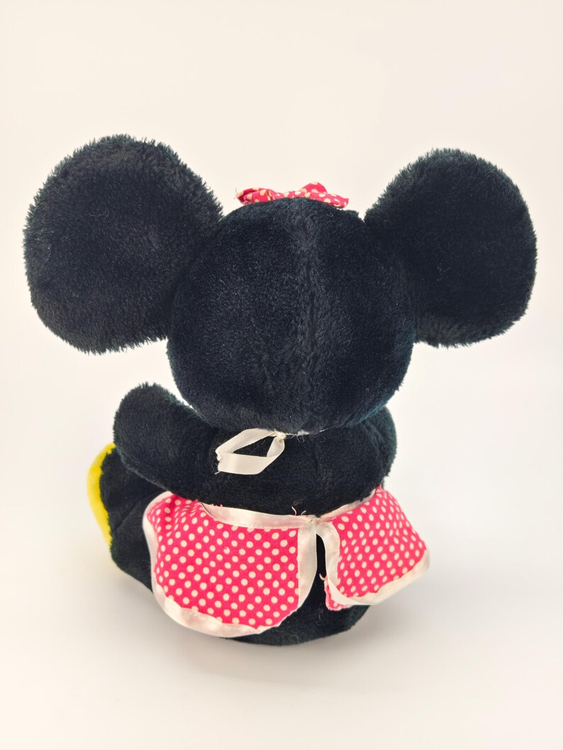 May include: A plush black mouse toy with large ears, a red polka dot bow, and a pink polka dot apron. The toy is sitting down, with its back facing the viewer. The apron has white trim.