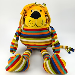 Puede incluir: Un león de peluche con cara y melena amarillas, y una nariz negra. El cuerpo, los brazos, las piernas y la cola están cubiertos de rayas horizontales de colores. El juguete tiene una expresión amigable y está diseñado para niños.
