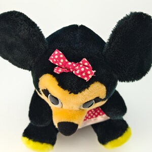 May include: A plush toy mouse with black fur, large black ears, and a yellow face. It has a red bow with white polka dots and yellow accents on its feet. The toy is a classic children's item.