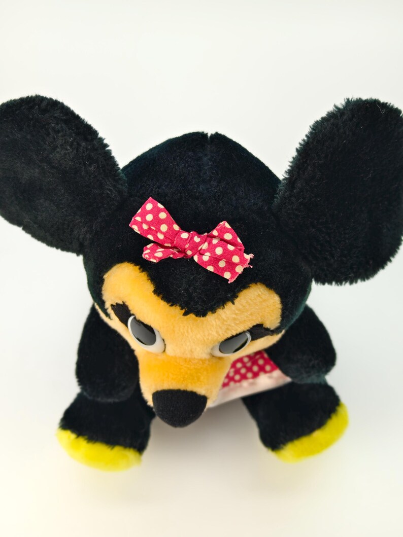 May include: A plush toy mouse with black fur, large black ears, and a yellow face. It has a red bow with white polka dots and yellow accents on its feet. The toy is a classic children's item.