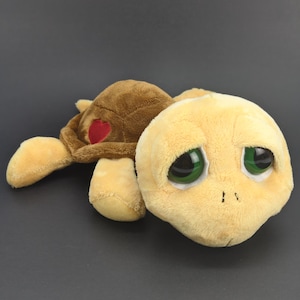 Pode incluir: Um brinquedo de pelúcia em forma de tartaruga, com cabeça e membros amarelo-claros e grandes olhos verdes. O casco é castanho com um pequeno remendo em forma de coração vermelho na lateral. O brinquedo tem uma textura macia e felpuda.