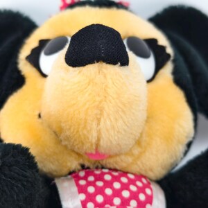May include: Close-up of a plush toy dog with a yellow face, black ears, and a black nose. The dog has large white eyes and a pink bow on its head. It is wearing a pink dress with white polka dots.