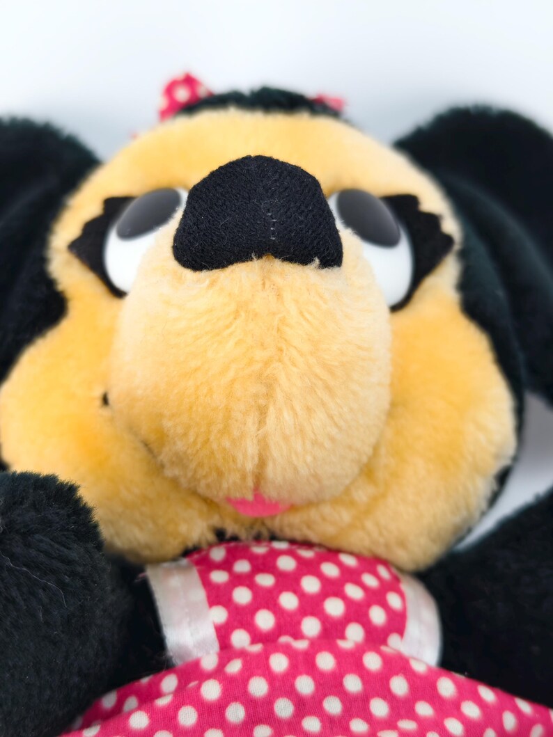 May include: Close-up of a plush toy dog with a yellow face, black ears, and a black nose. The dog has large white eyes and a pink bow on its head. It is wearing a pink dress with white polka dots.