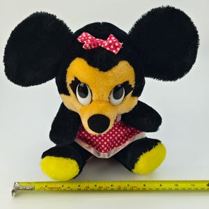 May include: A plush Minnie Mouse doll with large black ears, a yellow face, and a red polka-dot bow. The doll wears a red polka-dot dress and has yellow feet. A yellow tape measure is placed near the doll.