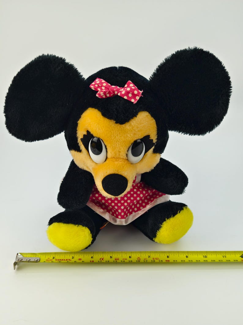 May include: A plush Minnie Mouse doll with large black ears, a yellow face, and a red polka-dot bow. The doll wears a red polka-dot dress and has yellow feet. A yellow tape measure is placed near the doll.