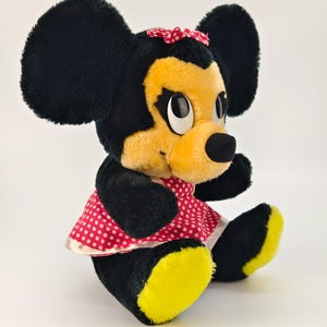 May include: A vintage plush toy of a cartoon mouse character. The mouse has large black ears, a yellow face, and a red and white polka dot dress. The toy is sitting with its legs extended and yellow feet.
