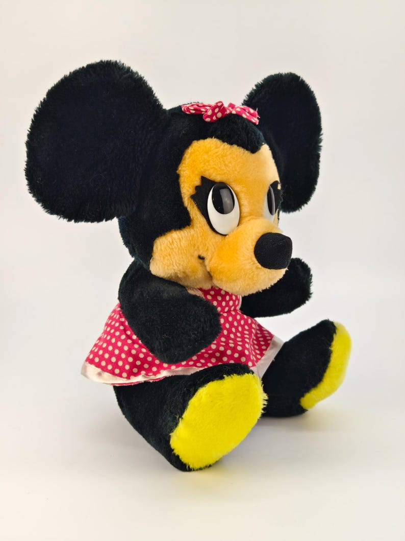 May include: A vintage plush toy of a cartoon mouse character. The mouse has large black ears, a yellow face, and a red and white polka dot dress. The toy is sitting with its legs extended and yellow feet.