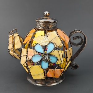 May include: A decorative teapot with a stained glass design. The teapot has a metal frame and handle, with yellow, orange, and blue glass pieces. A blue flower is in the center, and the lid is metal.