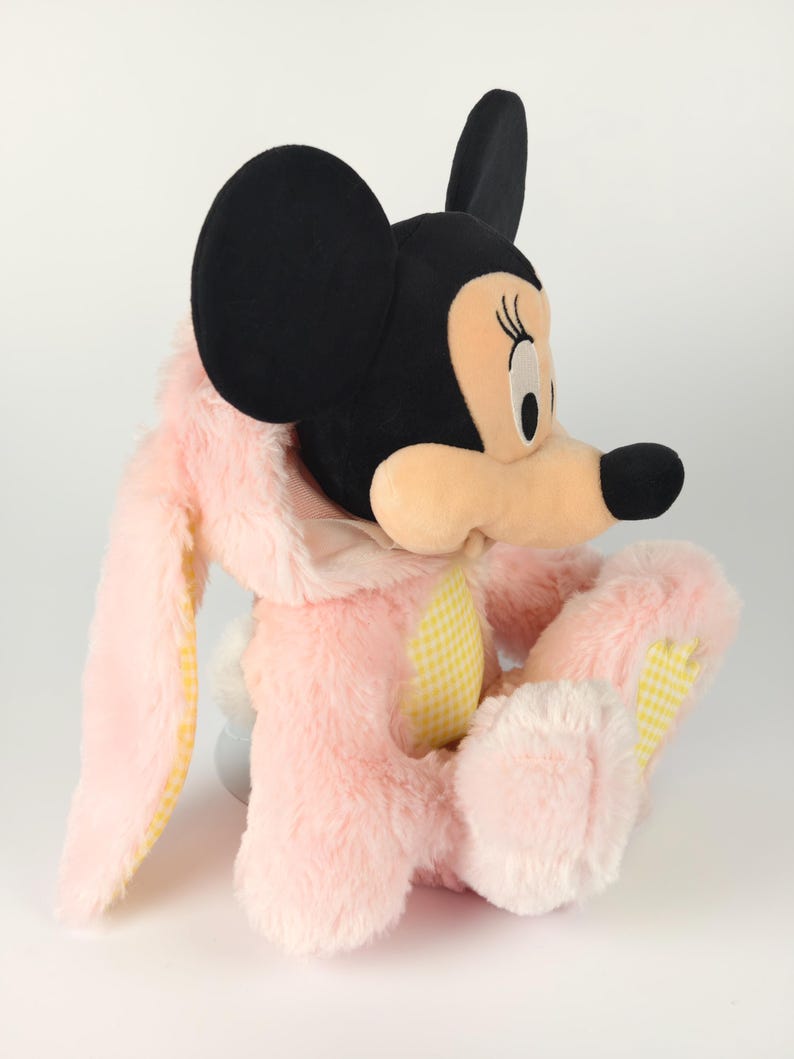 May include: A plush Minnie Mouse dressed as a bunny. The toy features a black head with large ears, a pink bunny suit with yellow gingham accents, and a white fluffy tail. The plush is soft and cuddly.
