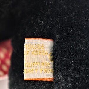 May include: Close-up of a black plush toy with a tag. The tag is rectangular with an orange border and features gold text that reads "HOUSE OF KOREA CLIPPINGS SHEY PRO". The plush toy has a soft, fuzzy texture.