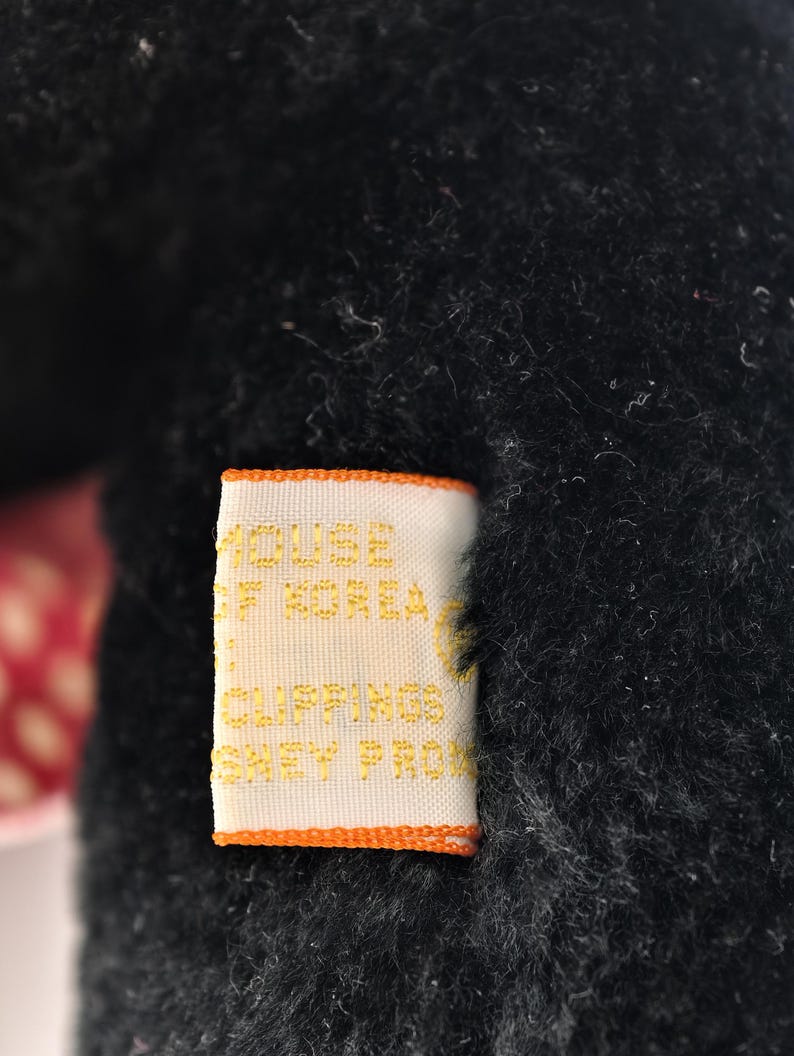 May include: Close-up of a black plush toy with a tag. The tag is rectangular with an orange border and features gold text that reads "HOUSE OF KOREA CLIPPINGS SHEY PRO". The plush toy has a soft, fuzzy texture.