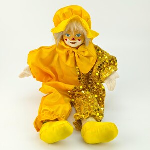 May include: A vintage clown doll in a yellow outfit, hat, and large shoes. The doll has a white face with blue eyes, red accents, and a large bow. One sleeve is decorated with gold sequins.