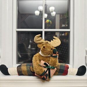 Può includere: Una decorazione a forma di alce di peluche con grandi corna, con un colletto bianco e pantaloni a quadri. L'alce tiene in mano un regalo avvolto con un nastro verde. L'alce è seduto su un davanzale.