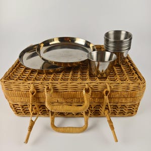 Puede incluir: Una cesta de picnic vintage hecha de mimbre tejido, con asa y cierres metálicos. La cesta contiene dos platos de acero inoxidable, una pila de pequeñas tazas de metal y dos tazas individuales de metal. La cesta es de color marrón claro.