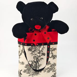 May include: A black velvet teddy bear with red lips emerges from a red and black gift bag. The bag has a black and white toile pattern, a black ribbon, and a handle. The bear has black button eyes.