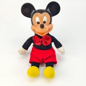 May include: A vintage Mickey Mouse soft toy. The toy has a black head with large ears, a tan face, and a red bow tie. It wears a black shirt, red shorts, and yellow shoes. The soft toy is set against a white background.