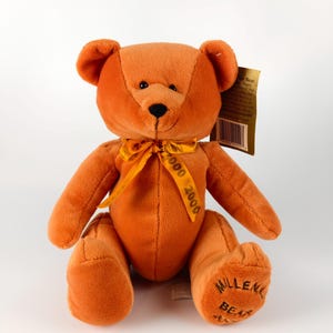 May include: An orange teddy bear with a yellow ribbon tied in a bow. The ribbon is printed with "2000". The bear has "Millennium Bear" printed on its foot. A gold tag is attached. The bear is made of plush material.