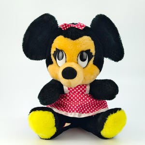 May include: A plush Minnie Mouse doll, featuring black fur, yellow face and feet, and a red polka dot dress. The doll has a red bow and large, expressive eyes. The toy is sitting upright against a white background.
