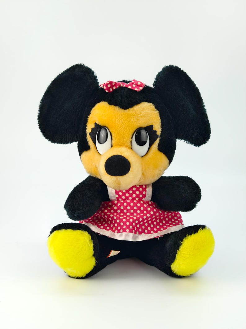 May include: A plush Minnie Mouse doll, featuring black fur, yellow face and feet, and a red polka dot dress. The doll has a red bow and large, expressive eyes. The toy is sitting upright against a white background.
