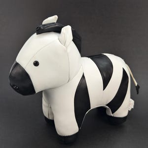 Puede incluir: Un juguete de cebra de peluche con un exterior de piel sintética lisa en blanco y negro. La cebra tiene el hocico, las orejas, la crin y los cascos de color negro. Se sostiene sobre cuatro patas y tiene una pequeña cola con un mechón negro.