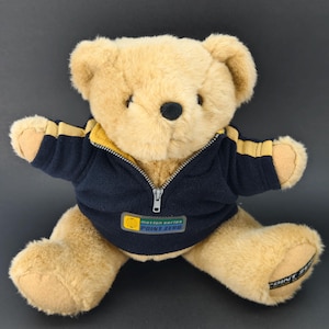 May include: A light brown teddy bear wearing a navy blue jacket with yellow stripes and a zip. The jacket has a small logo that reads "motion series Point Zero". The bear is sitting with its arms and legs spread.
