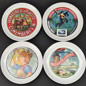 Peut inclure: Quatre assiettes en céramique blanche avec des motifs vintage de fromages Camembert. Chaque assiette présente une illustration différente, dont une vache, une femme et un moulin à vent, avec du texte en français. Les assiettes sont disposées sur une surface sombre.
