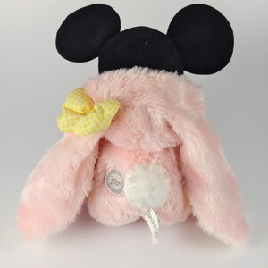 May include: A pink plush toy with black mouse ears and a yellow gingham bow. The toy has long, floppy ears and a white pom-pom tail. A Disney Store tag is visible.