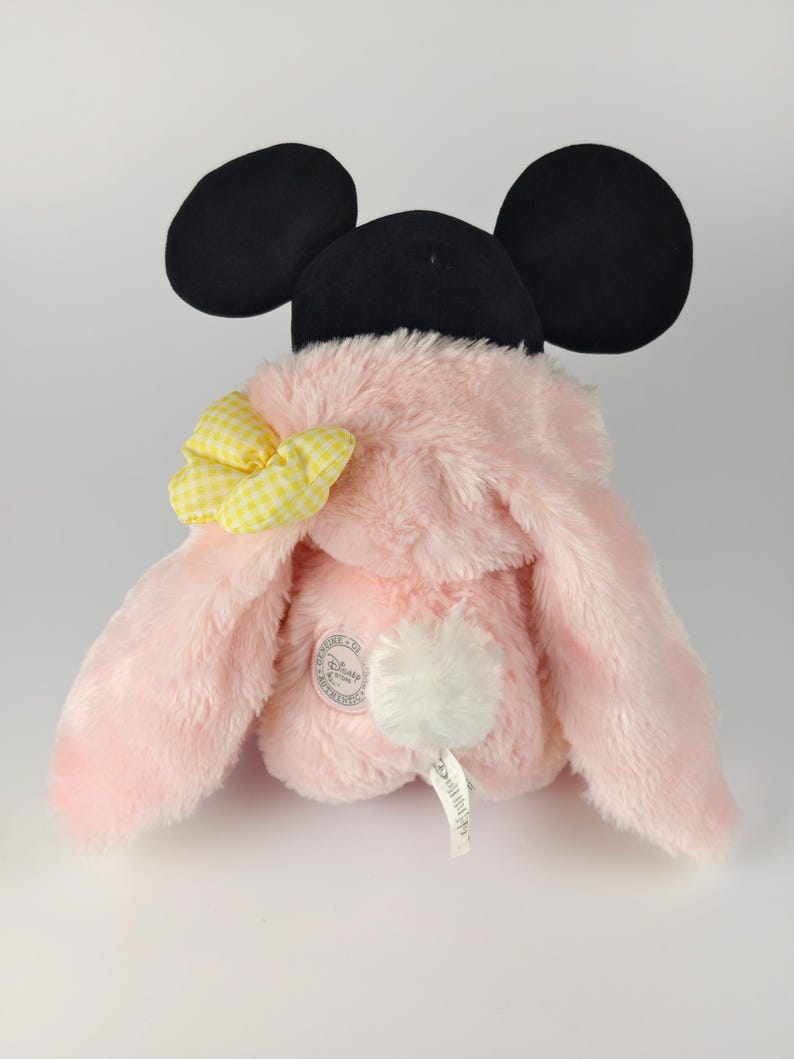 May include: A pink plush toy with black mouse ears and a yellow gingham bow. The toy has long, floppy ears and a white pom-pom tail. A Disney Store tag is visible.