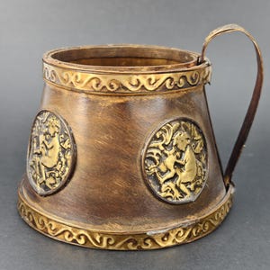 Peut inclure: Une chope en métal de couleur bronze avec une anse. La chope présente deux motifs circulaires en relief avec des figures dorées et une bordure dorée ornée en haut et en bas. La poignée est également dorée.