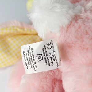 May include: A close-up of a pink plush toy with white accents. A tag is attached, displaying text including "ALL NEW MATERIALS" and "MADE IN VIETNAM". The toy has a yellow and white checkered pattern.