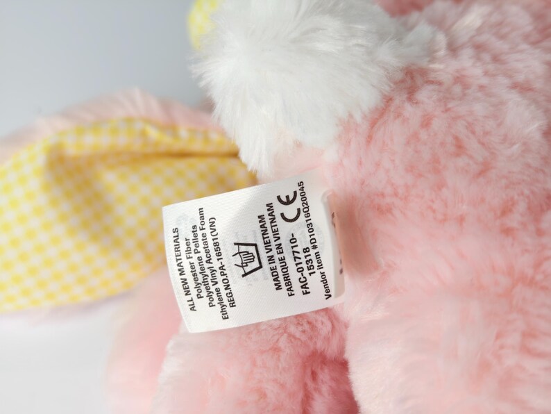 May include: A close-up of a pink plush toy with white accents. A tag is attached, displaying text including "ALL NEW MATERIALS" and "MADE IN VIETNAM". The toy has a yellow and white checkered pattern.