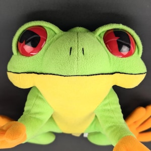 Op de afbeelding: Een pluche kikker met grote rode en zwarte ogen, een groene body, gele buik en oranje voeten. De kikker is gemaakt van zachte stof en heeft een vriendelijke uitdrukking. Het speelgoed is ontworpen om op een boomkikker te lijken.