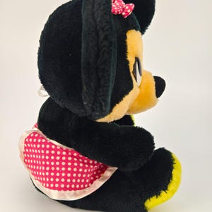 May include: A plush Minnie Mouse doll, featuring black fur, a yellow face and feet, and a pink polka-dot apron. A small pink bow with white polka dots adorns the top of the head. The doll is in a seated position.