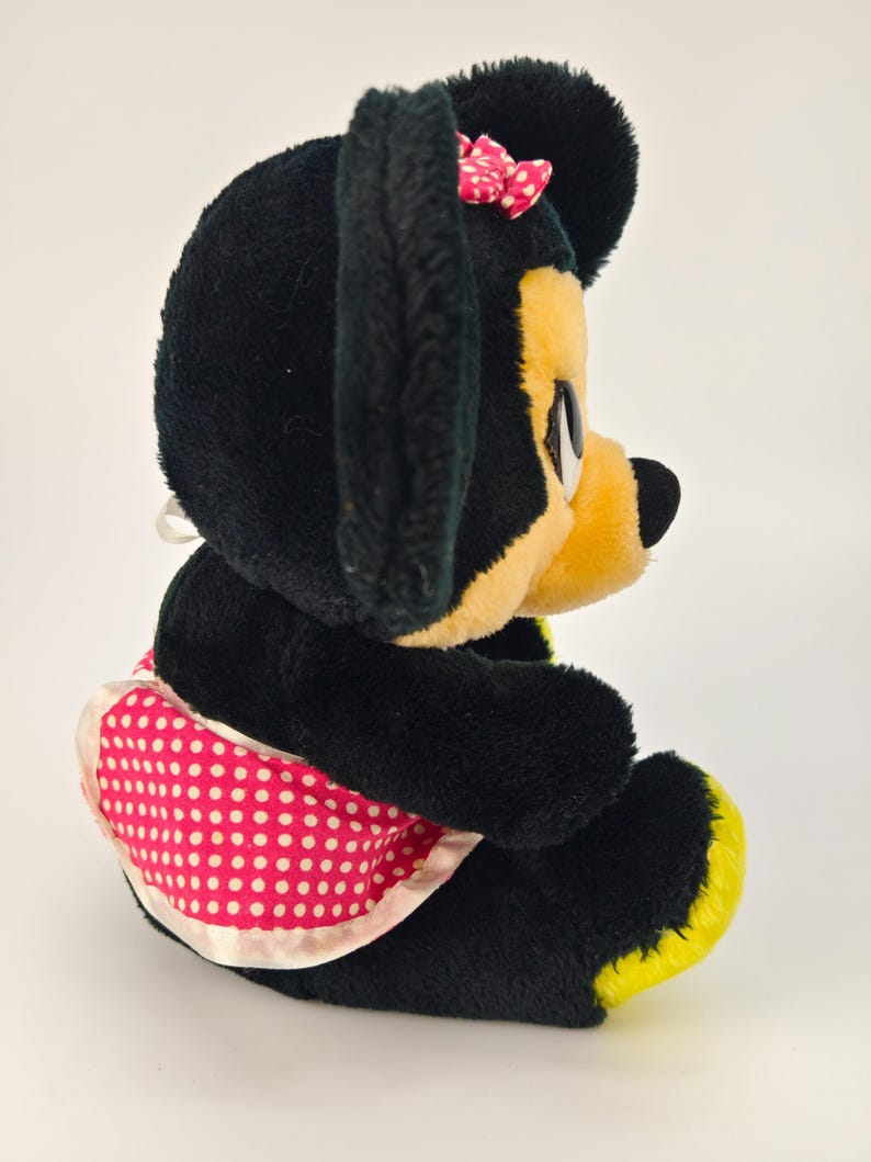 May include: A plush Minnie Mouse doll, featuring black fur, a yellow face and feet, and a pink polka-dot apron. A small pink bow with white polka dots adorns the top of the head. The doll is in a seated position.