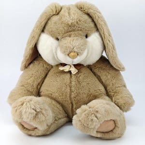 May include: A soft, plush bunny rabbit toy in shades of beige and cream. The bunny has long, floppy ears, a cream-coloured face, and a small bow around its neck. The toy is made of soft, fluffy material, perfect for a child.