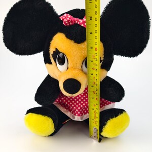 May include: A plush Minnie Mouse doll, featuring black fur, yellow face and feet, and a red polka-dot dress. The doll has a pink bow and is approximately 11 inches tall, as measured by a yellow tape measure.